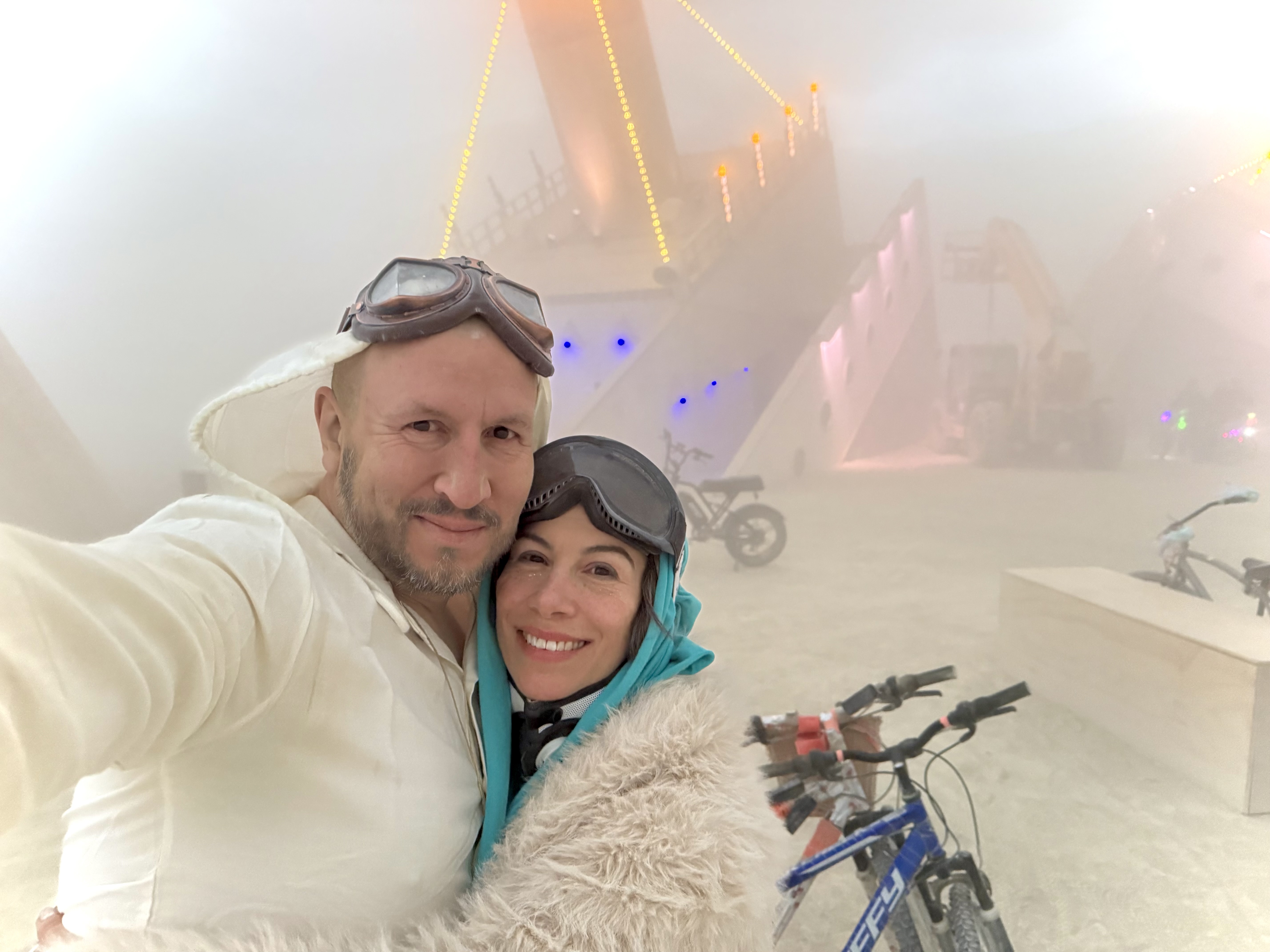 Alexander Torrenegra and Tania Zapata in white desert outfits with goggles at Burning Man